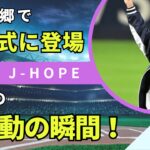 BTSのJ-HOPE（ホビ）が光州の始球式に登場し、地元ファンから大歓声を浴びる様子。ホビは紫のグローブを持ち、野球場のマウンドで堂々とした姿を見せています。